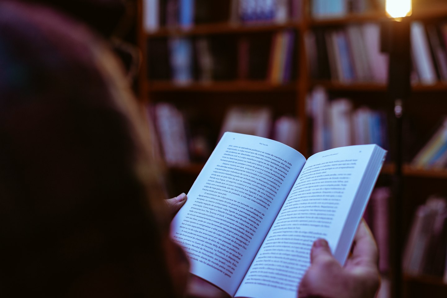 Persona leyendo un libro abierto con estanterías borrosas detrás.